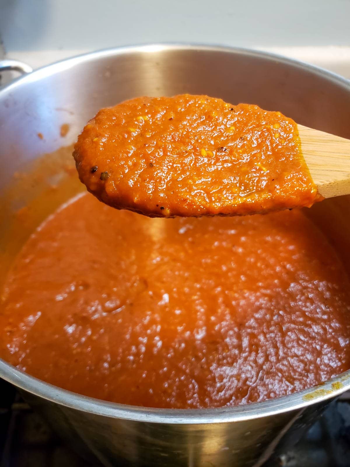 A close up image of the finished roasted tomato sauce on a wooden spoon hovering over a large stainless steel pot full of sauce inthe background
