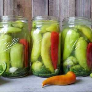 Three jars of pickled peppers in a row, full of mostly yellow-green banana peppers and few hot red ember chilis