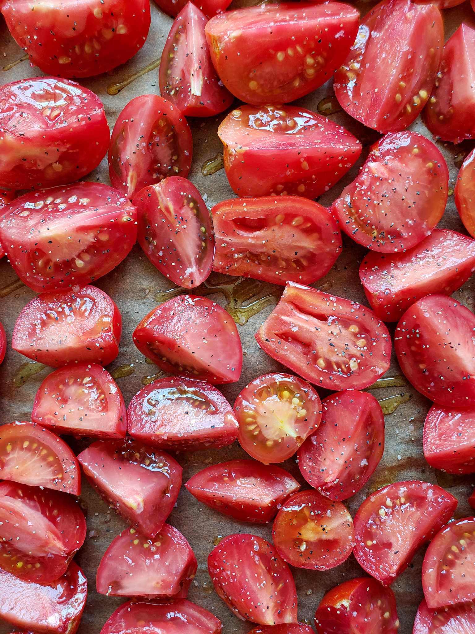 A closeup image of the halved and quartered red tomatoes after they have been drizzled with olive oil and sprinkled with sea salt and black pepper. They are now ready to go into the oven.
