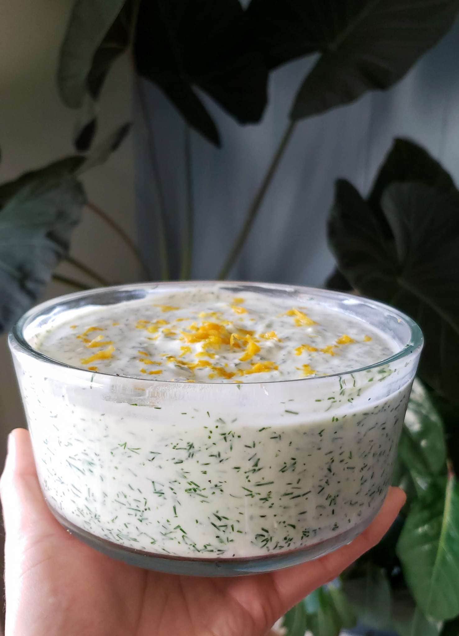 A hand is holding a round glass bowl full of yogurt dipping sauce or dressing with dill, lemon, and garlic. There is chopped dill mixed throughout the yogurt while lemon zest adorns the top of the sauce.