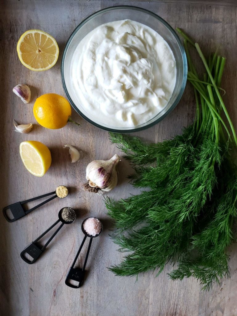 The ingredients for the yogurt sauce covers a wooden surface. A  bowl of yogurt, two lemons, one that is sliced in half, garlic cloves, a bulb of garlic, three measuring spoons each with salt, pepper, and onion powder, and a bunch of dill are laid out in an equally spaced arrangement.