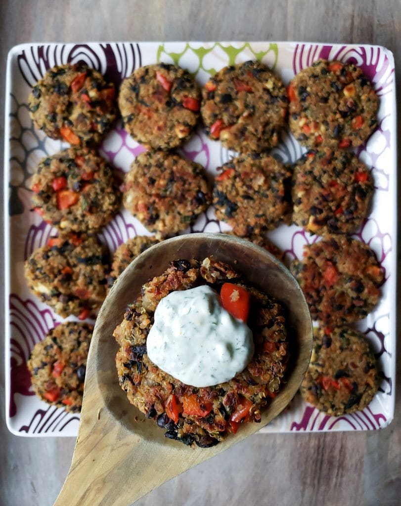 A close up of a wooden ladle with a quinoa burger in the center of it, you can see there is also black bean and red bell pepper in the quinoa pattie as well. There is a half dollar sized drop of yogurt dill sauce in the center of the patty, showing brightly against the darker veggie patty. Below the featured patty there is a tray full of quinoa burgers lined up in a neat grid of 4x4.