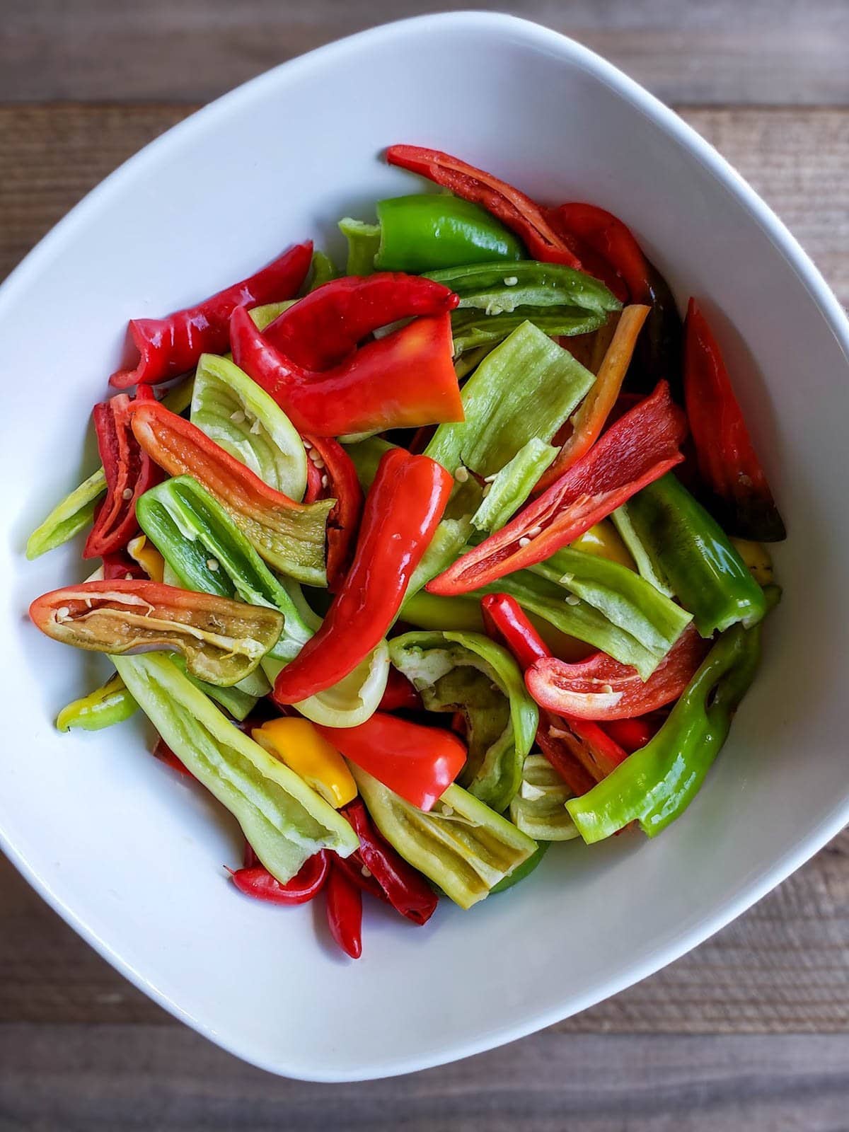 A white ceramic bowl full of red, green and yellow chili peppers that have all been sliced in half and cleaned of their membrane and seeds. 