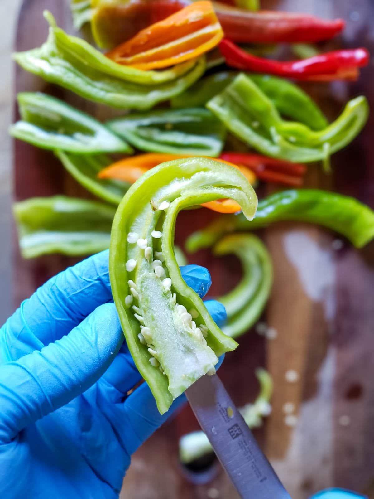 A gloved hand holding a chili pepper that has been sliced in half, with a knife being used to cut away and remove the inner membrane and seeds 