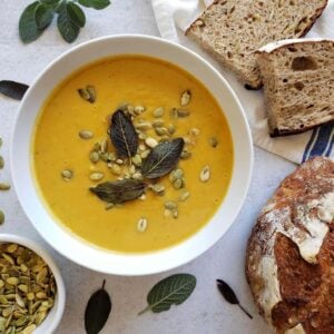 a bowl of butternut squash soup with sliced bread, pumpkin seeds, and sage leaves scattered around it