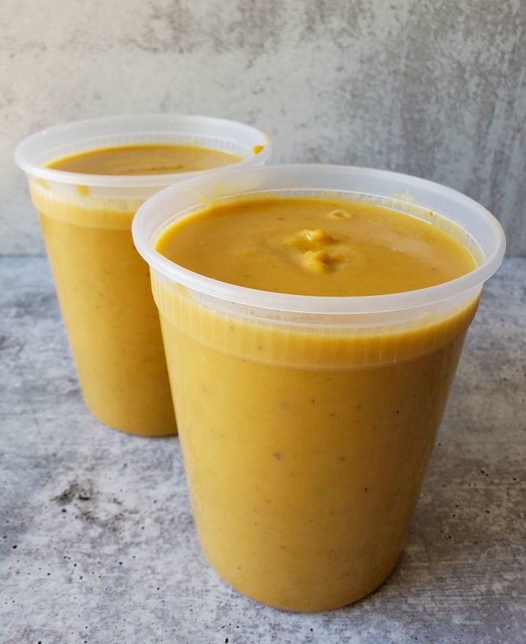 Two plastic BPA free quart jars sit full to the brim with the roasted butternut squash soup. The background is a washed concrete surface, and the soup stands out with its bright golden yellow color. These can now be refrigerated or frozen for future use.