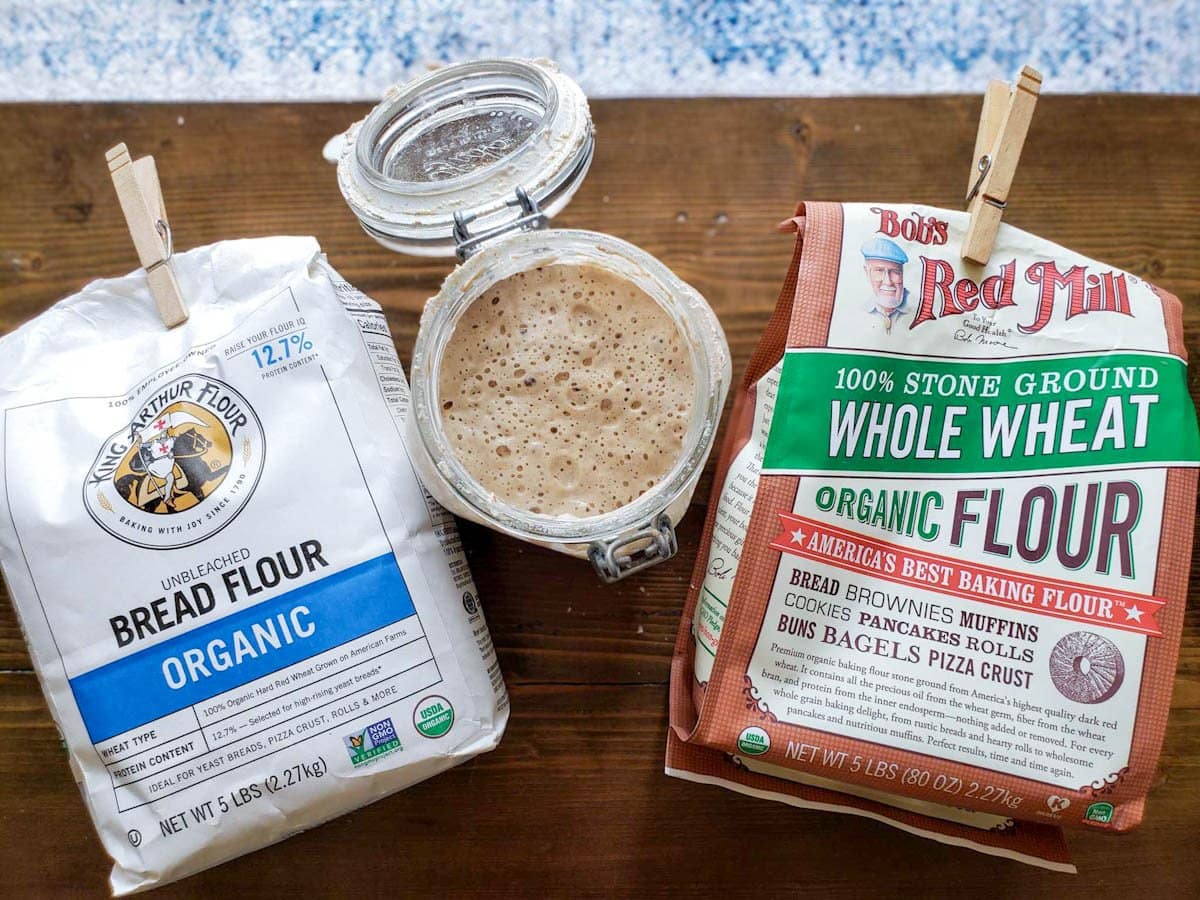 Looking down at a table with a bag of King Arthur Flour Organic Bread Flour facing upwards next to a glass container that contains bubbly sourdough starter, on the other side of the jar is a bag of Bob's Red Mill Organic Whole Wheat Flour. They are all sitting on a dark barn wood surface.