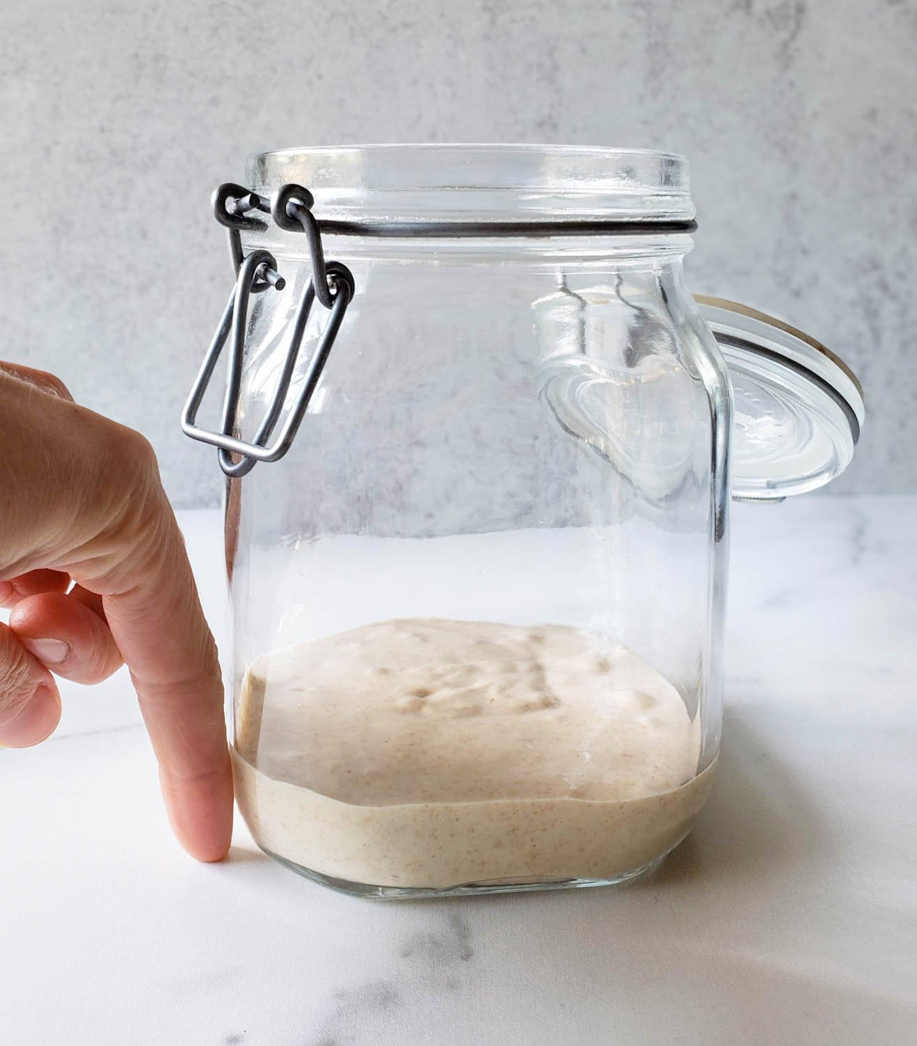 A close up of a glass flip top glass container from the side with the starter only filling the container about one inch deep. A finger is pointed downward, touching the ground next to the container showing the starter depth in relation to a finger.