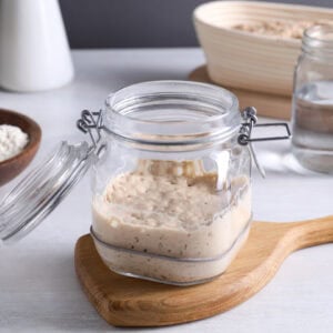 A glass jar with a flip top lid is halfway full of sourdough starter. Flour, water, and a banneton are in the background.