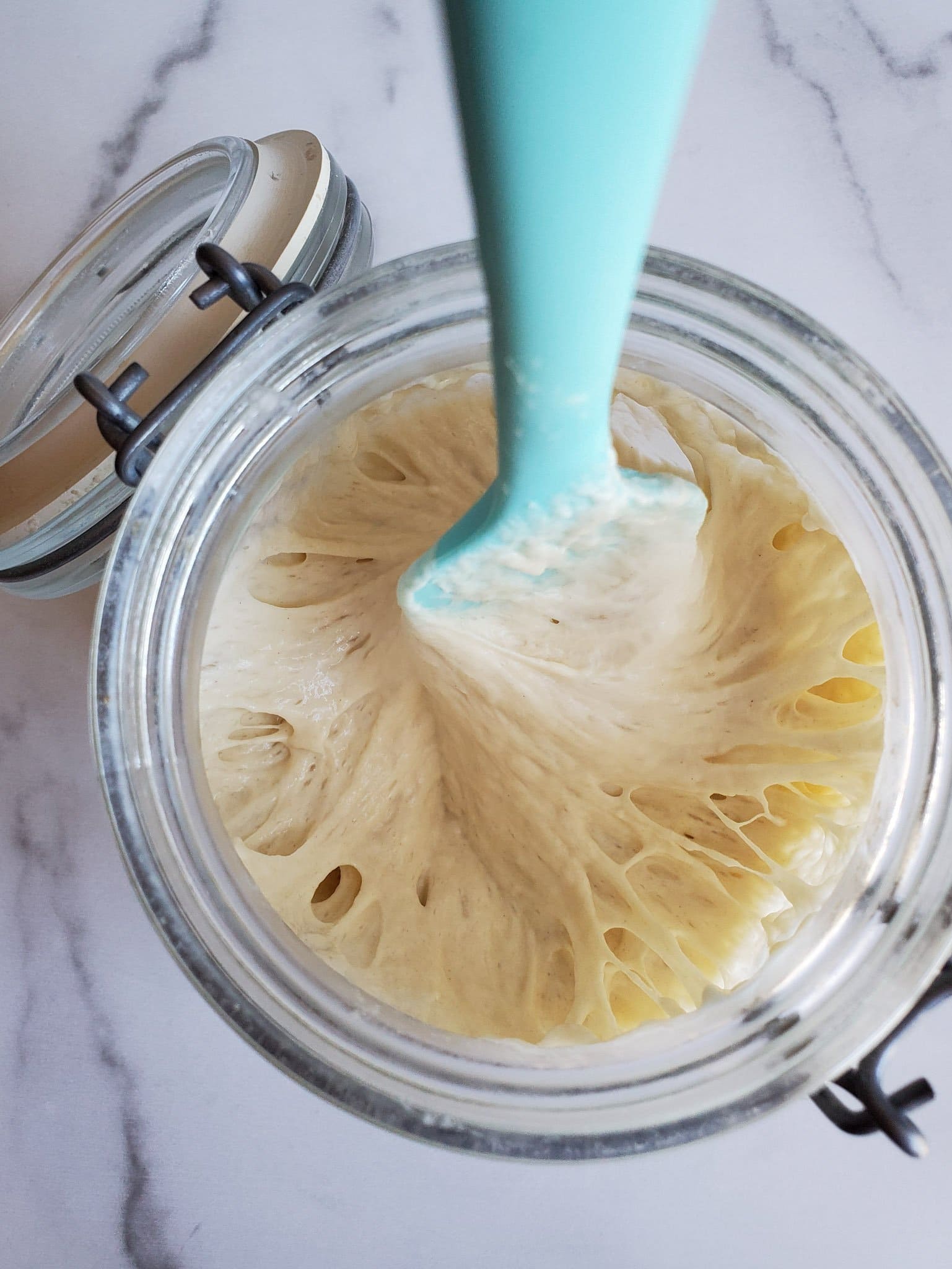 A blue silicone spatula is stirring sourdough starter inside of a glass jar with a flip top lid.
