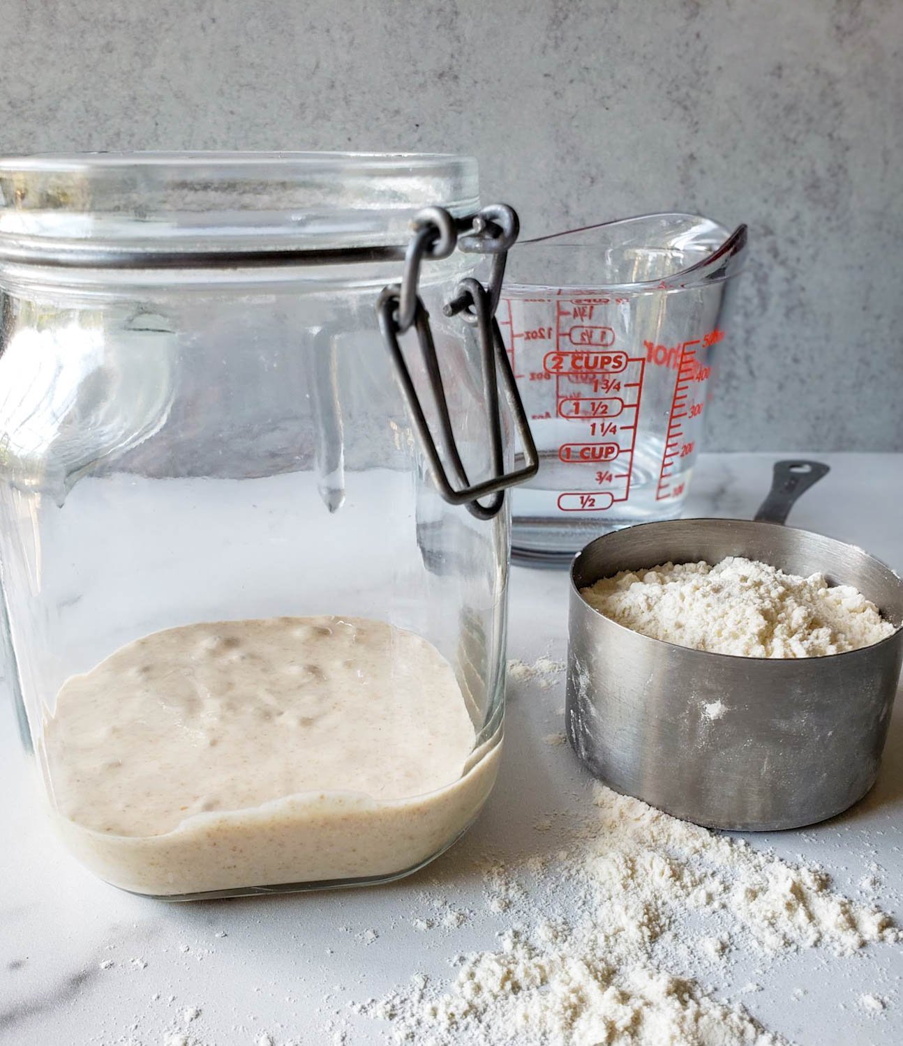 A large glass jar with a small amount of flour along with a metal measuring cup of flour and a liquid glass measuring cup are arranged near on another.
