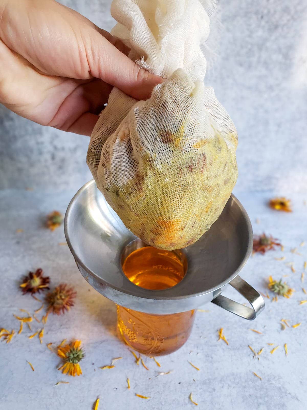 A section of cheesecloth filled with dried flowers is held above a metal funnel that is place on top of a pint mason jar filled with oil. Dried flowers are scattered about the surrounding area.
