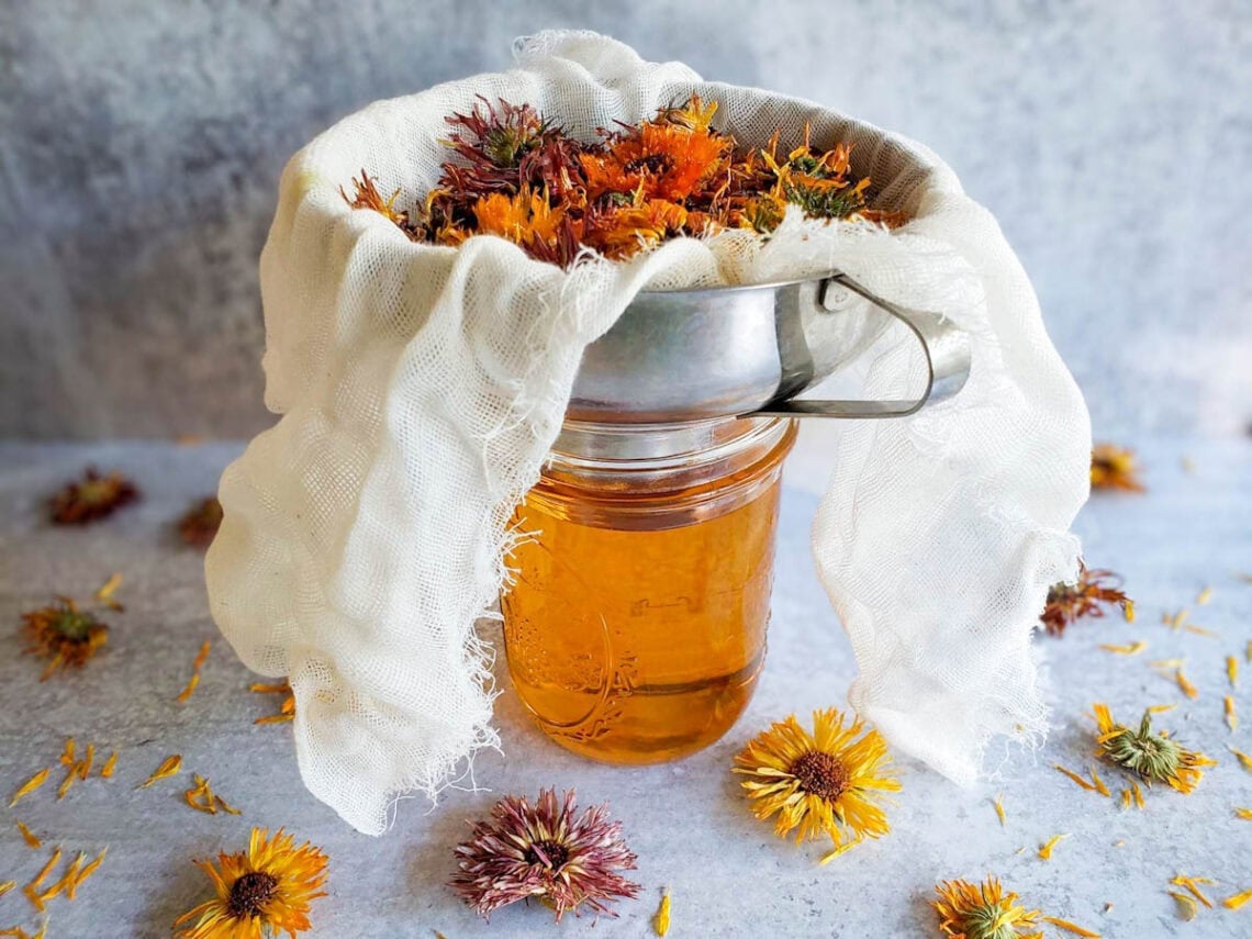 A pint mason jar is fully of calendula oil, a metal canning funnel sits on top of the jar which is lined with cheesecloth that contains dried calendula flowers.