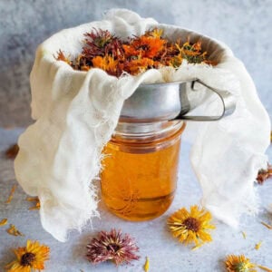 A pint mason jar is fully of calendula oil, a metal canning funnel sits on top of the jar which is lined with cheesecloth that contains dried calendula flowers.