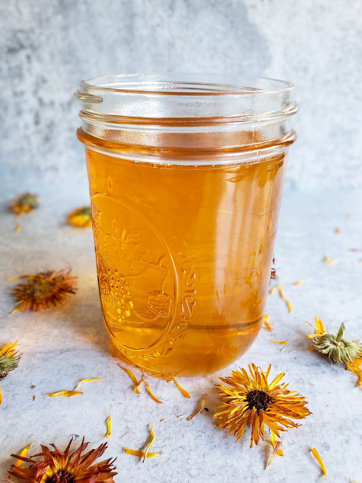 A glass mason pint jar full of calendula oil, many dried flowers garnish the surrounding area.