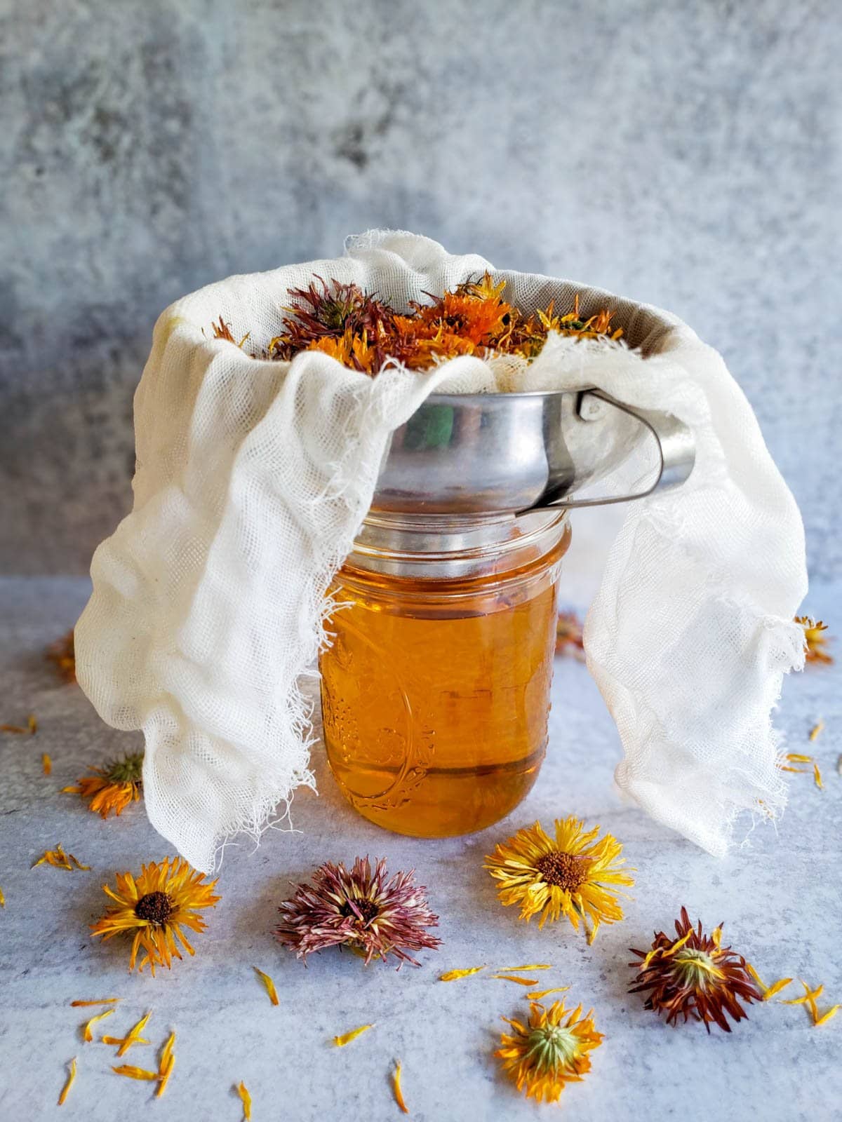 Dried flowers are inside cheesecloth that is placed over the top of a metal funnel that is sitting on a pint jar.