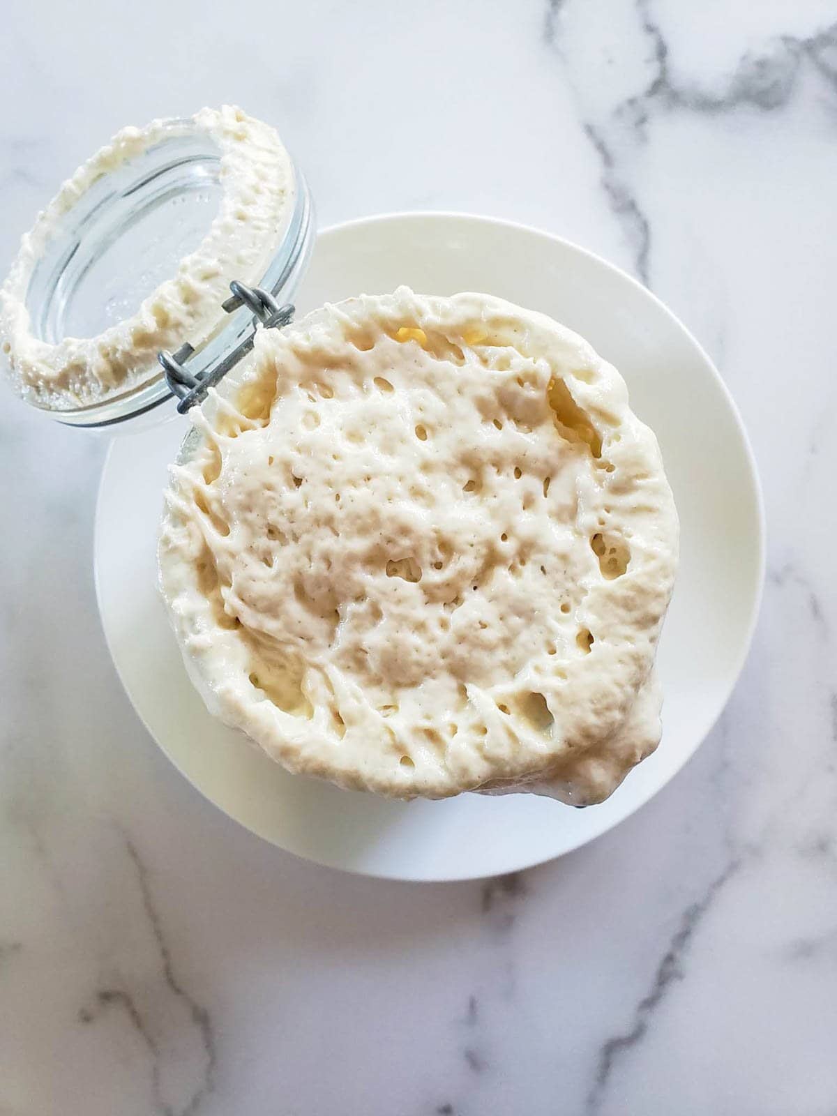 Pillowy sourdough starter is bubbling out of the top of a glass jar with a flip top lid.