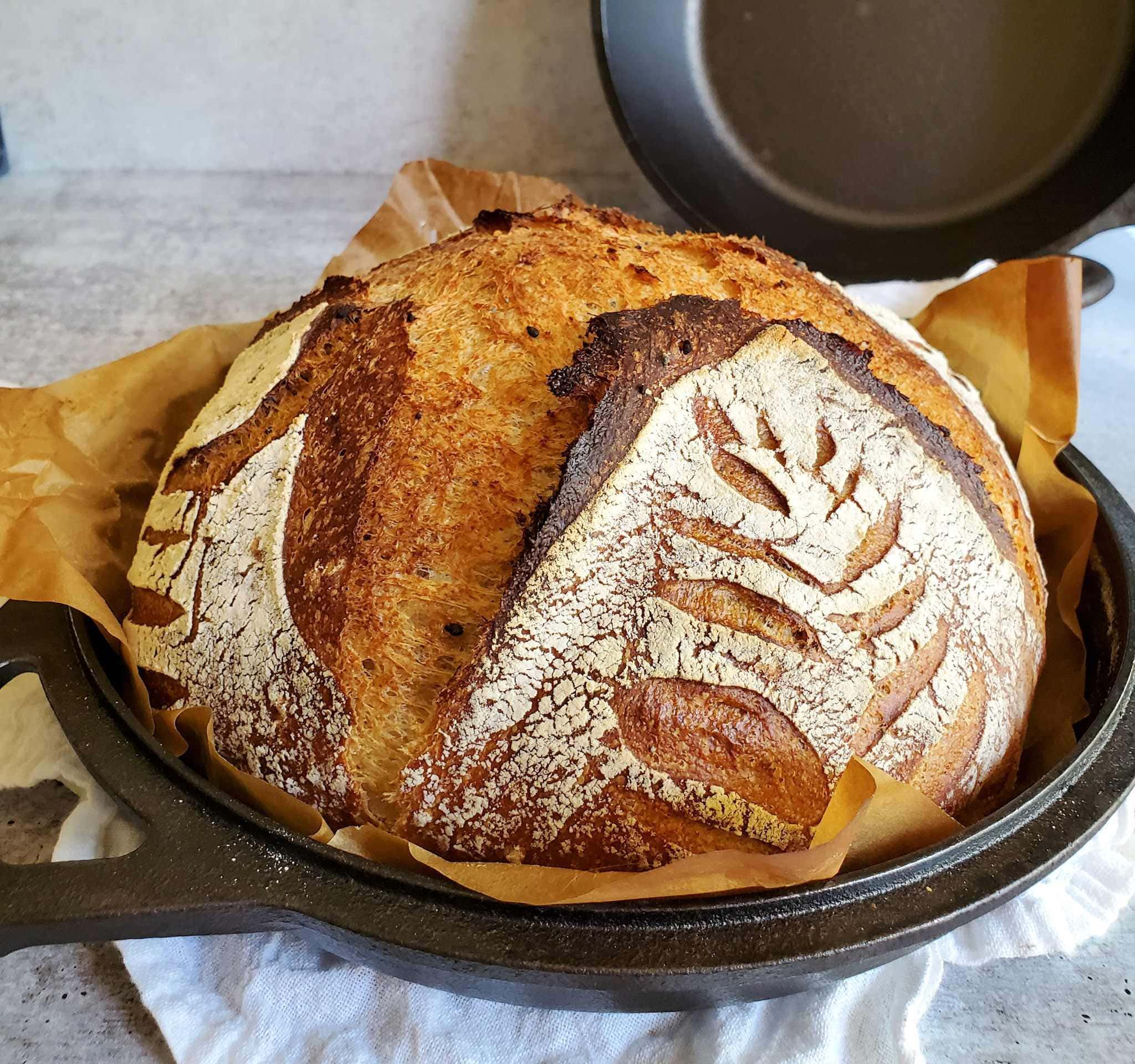 A round loaf of baked sourdough bread sits inside of a cast iron combo cooker after being removed from the oven. It has shallow leaf-like designs that have been carved into the sides with a bread lame.