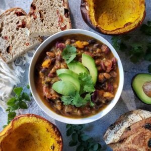 A bowl full of pumpkin chili garnished with avocado slices and cilantro. Surrounding the bowl are a few slices of sourdough bread, roasted halves of whole pumpkin, and a half of an avocado.