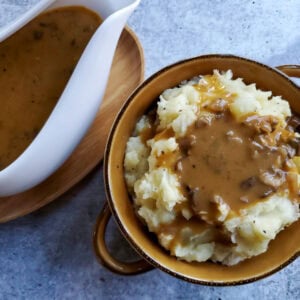 looking down on a brown bowl with handles full of mashed potatoes covered in chunky mushroom gravy, with a gravy boat set off to the side