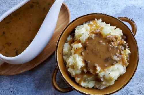 looking down on a brown bowl with handles full of mashed potatoes covered in chunky mushroom gravy, with a gravy boat set off to the side