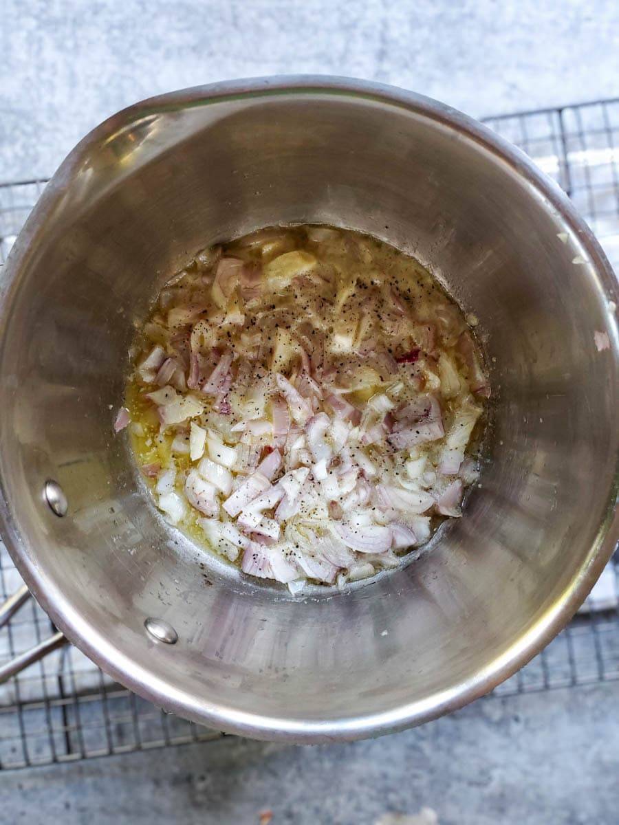 A close up inside a saucepan with olive oil, diced shallots (onions) and garlic in the bottom of the plan (still raw but cooking), with visible salt and pepper sprinkled over.