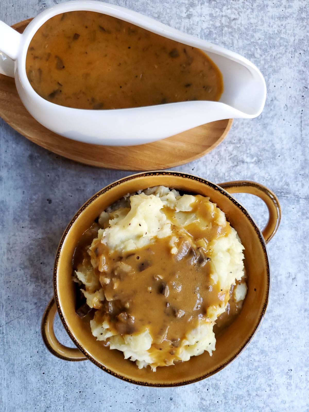 A bowl of mashed potatoes topped with mushroom gravy, with a full gravy boat sitting off to the side