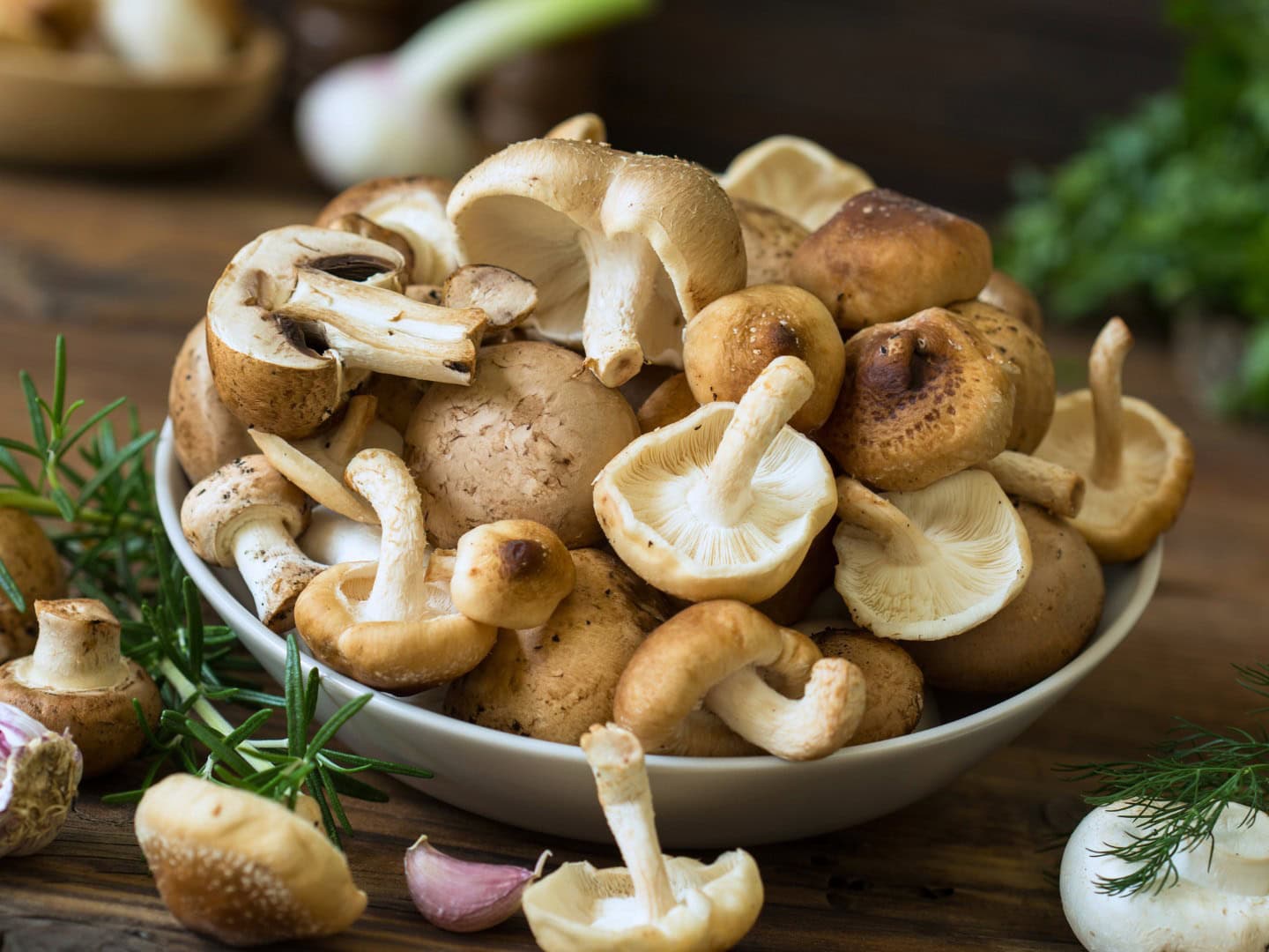 A bowl of raw, whole mushrooms laying on a wooden table with herbs and onions around it