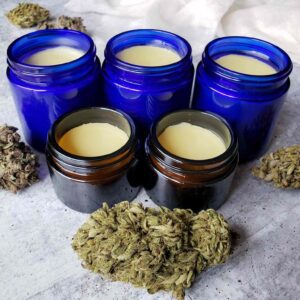 Five small glass jars are each filled with cannabis salve. Cannabis flowers surround the jars.