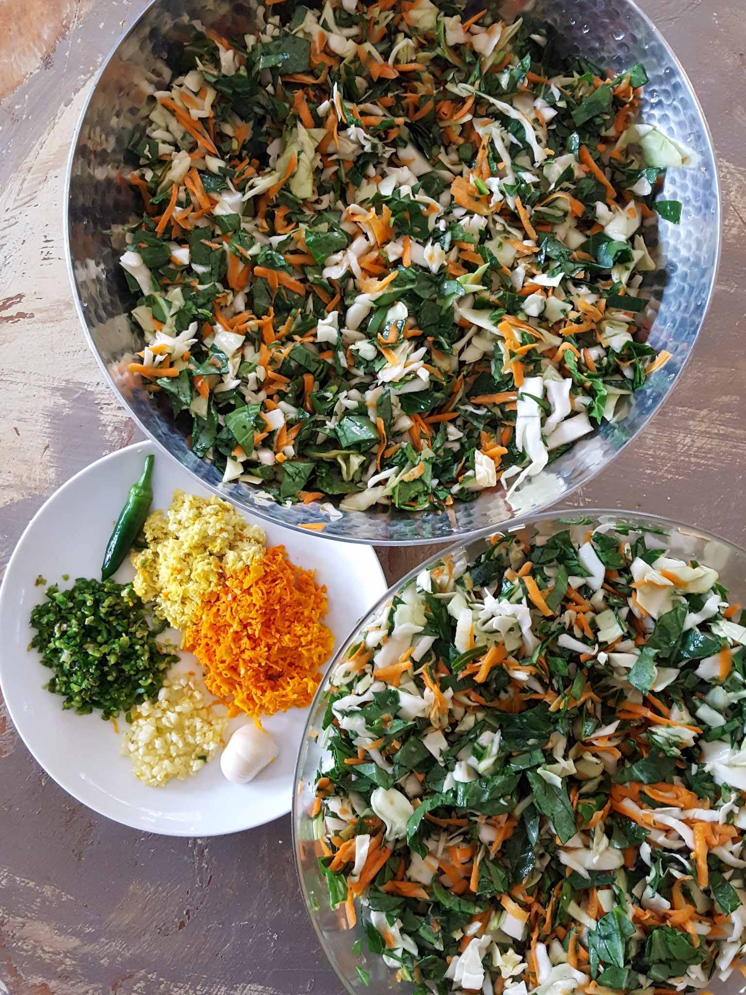 Two bowls of various chopped greens and grated carrot are shown. There is a small white plate next to the bowls that contains the seasonings for the kraut. Chopped fresh turmeric, chopped fresh ginger, chopped fresh garlic, and chopped fresh chili peppers are taking up their portion of space on the plate.