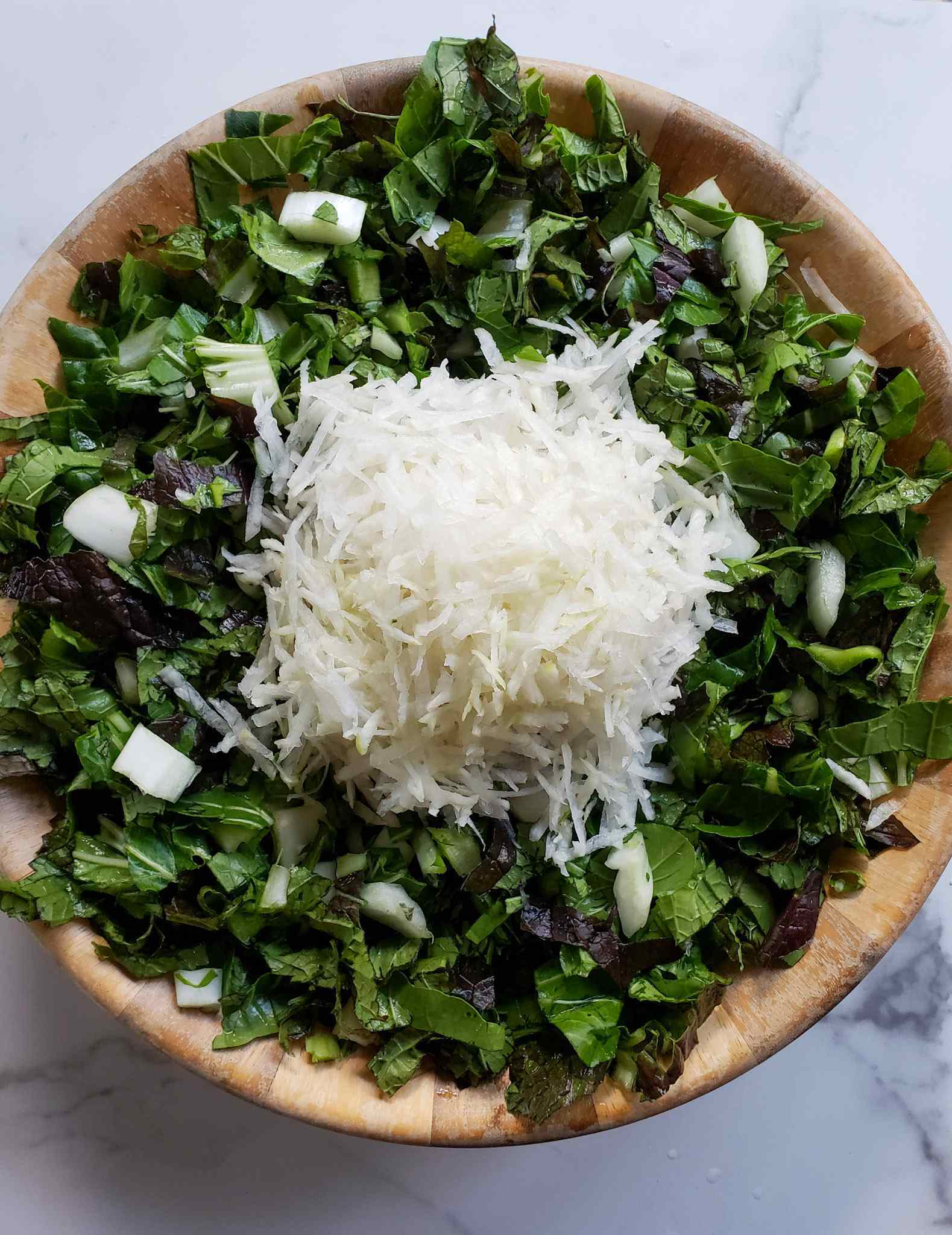 A large wooden bowl is shown full of chopped bok choy and red mustard greens. A pile of grated daikon radish is sitting in the middle of the chopped greens. 