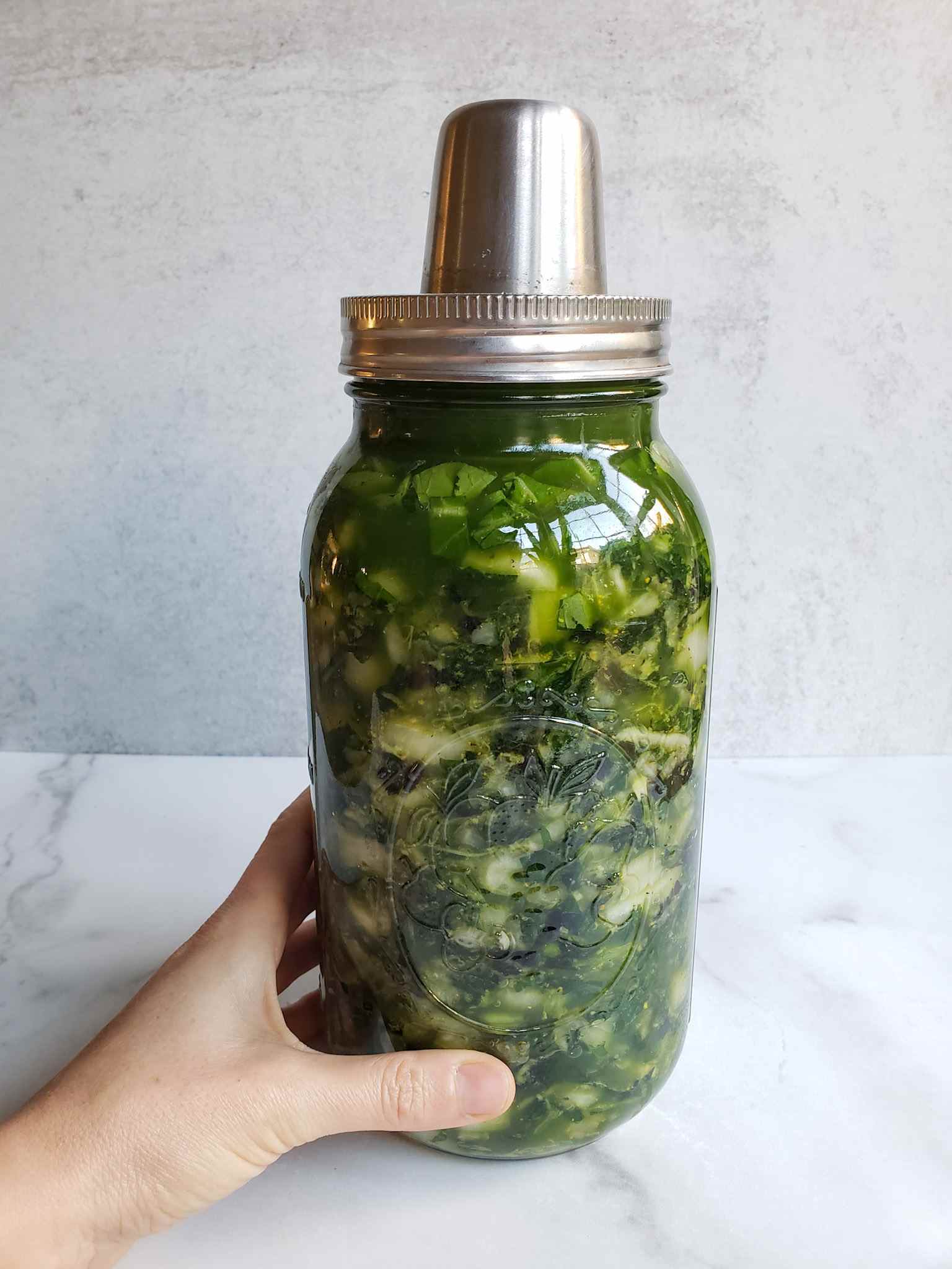 A half gallon mason jar full of fermented green kraut is shown with an air lock ferment lid attached on top.  A hand is partially holding the jar, as if to steady it to take a clear picture. The jar is full of greens and there is a beautiful green brine that has formed, mostly visible in the top third of the jar. 