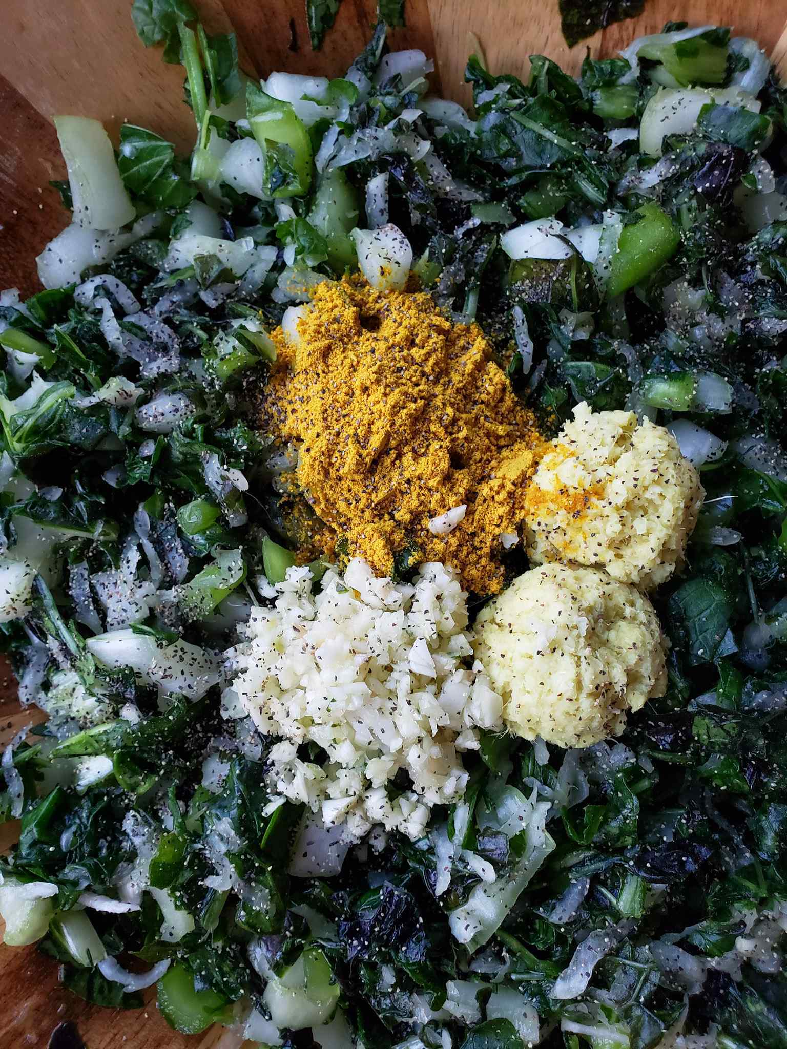 A close up image of chopped greens with two tablespoons of fresh ginger, one tablespoon of fresh garlic, one tablespoon of turmeric powder, and a sprinkle of black pepper  sitting neatly atop the greens. 