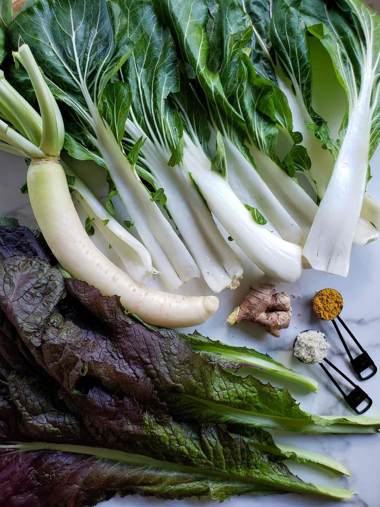 Leaves of bok choy, red mustard greens, a daikon radish, a chunk of ginger, and a tablespoon each of celtic sea salt and turmeric powder are displayed on top of a marble surface. These are the ingredients of a green kraut.  