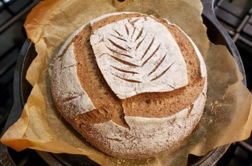 a close up of a freshly baked gluten free sourdough bread loaf still inside the cast iron dutch oven, with a golden brown crust and wheat leaf pattern carved into the top white flour