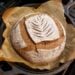 a close up of a freshly baked gluten free sourdough bread loaf still inside the cast iron dutch oven, with a golden brown crust and wheat leaf pattern carved into the top white flour