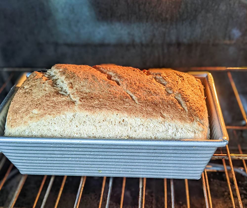 a close up of a loaf of bread still inside the oven in a classic rectangular 1 lb bread pan loaf. The bread has risen above the rim of the pan and has a golden brown crust