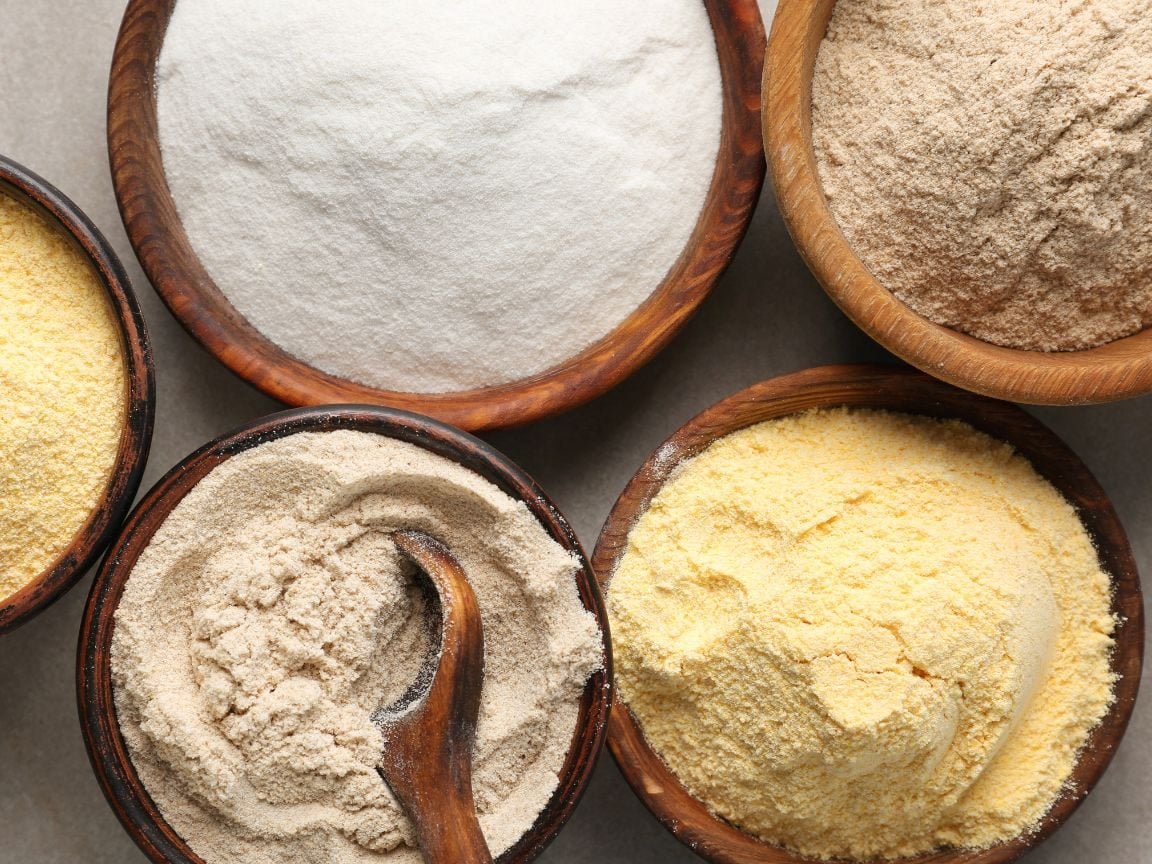 A close up looking down on 5 wooden bowls full of different gluten free flours, some are more brown, tan, white or yellow in color.