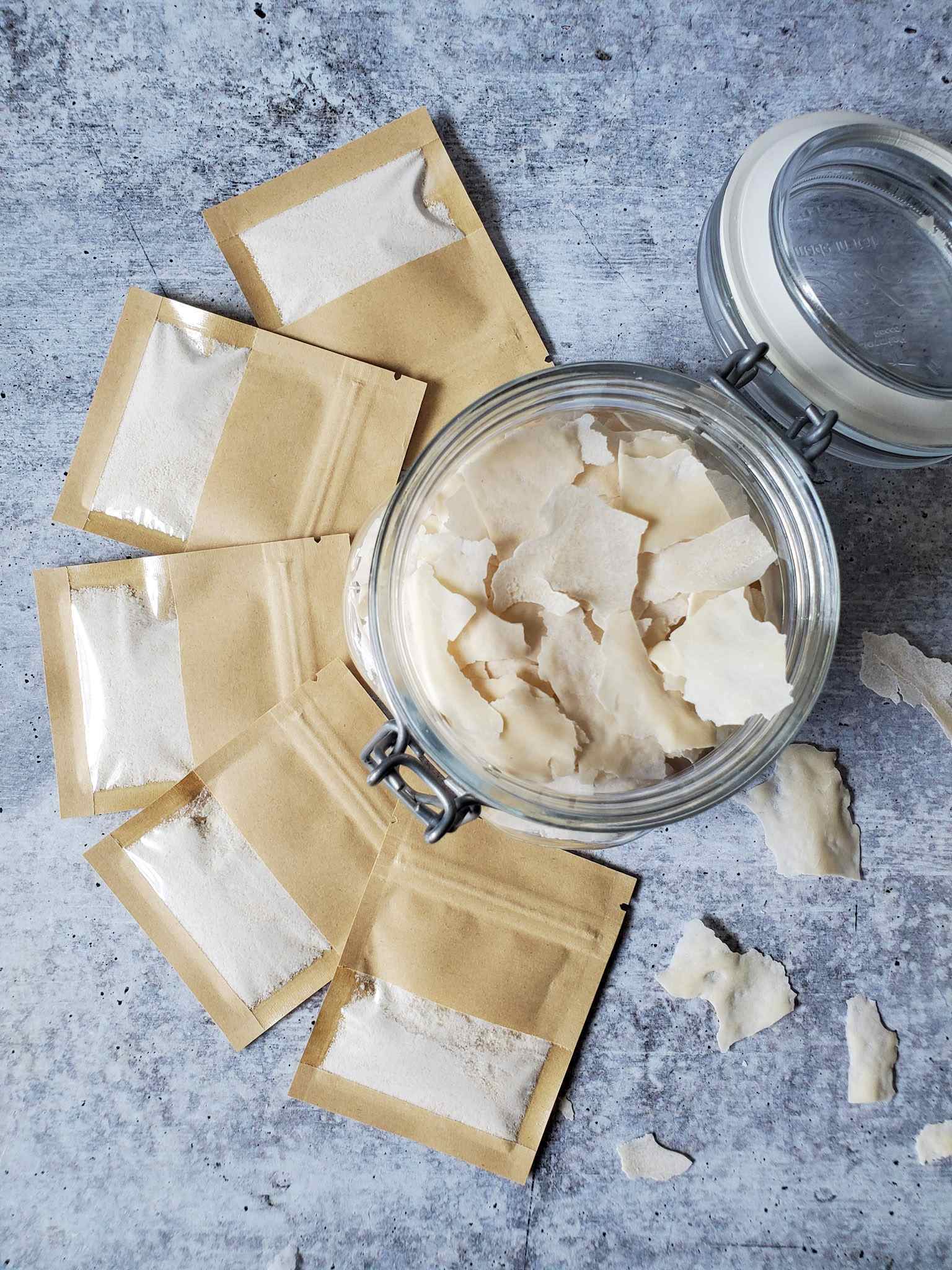 A small glass jar is full of dry flakes of sourdough starter, like small thin white chips, with several small brown packets or envelopes of dried sourdough starter sitting around it