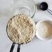 a close up of a tablespoon full of dry sourdough stater powder, with a jar of water and flour in the backgroun