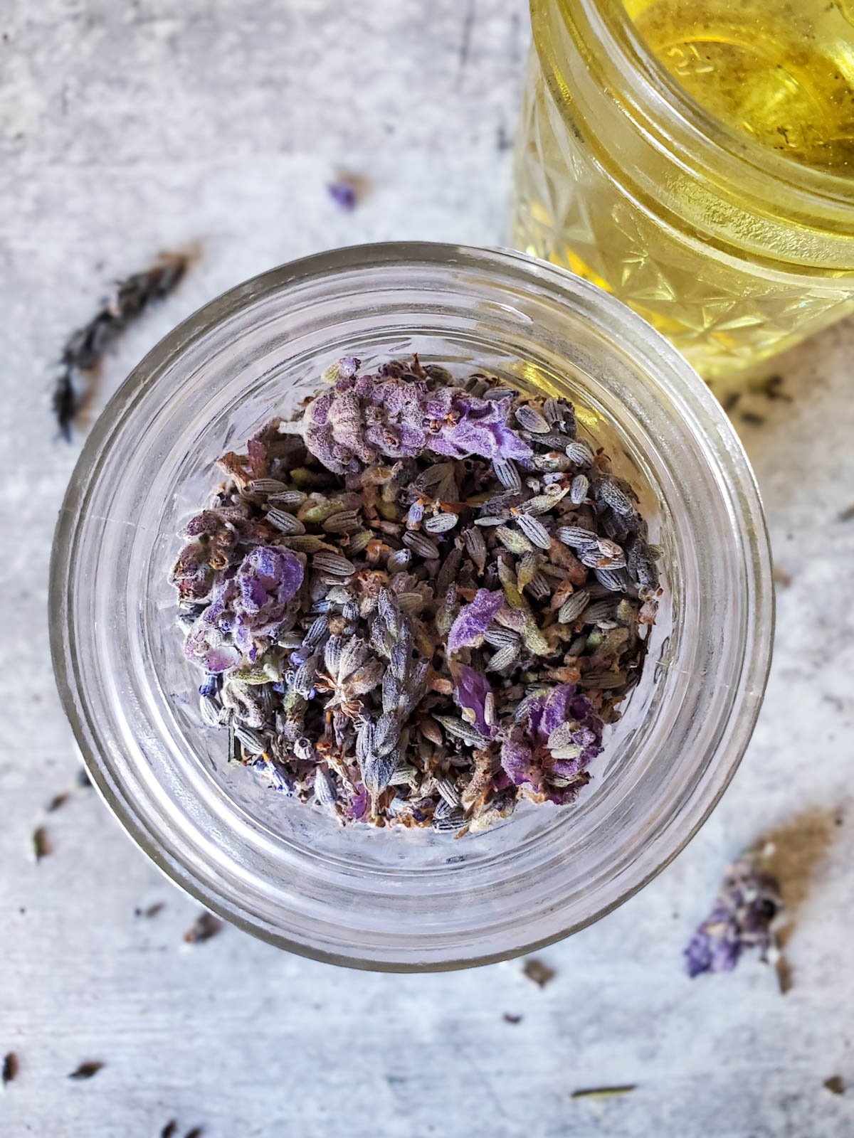 A small mason jar full of dehydrated lavender flowers. A jar of oil is nearby with a few dried flowers scattered about the area. 