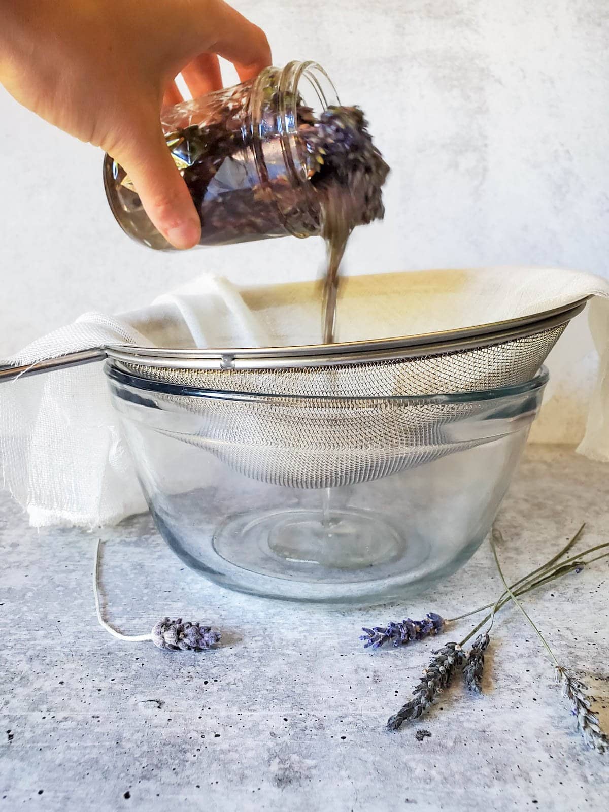A jar of infused oil is being poured through a strainer that is lined with cheesecloth into a glass bowl. 