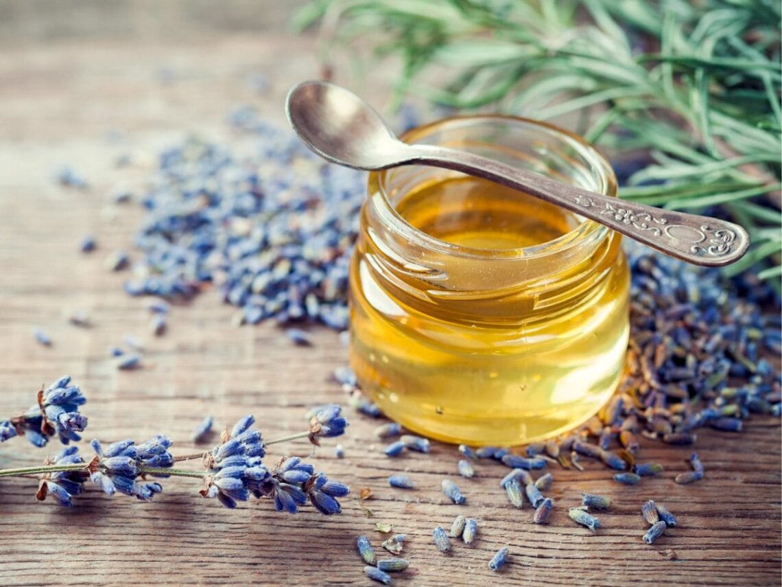 A small jar of lavender oil with tiny lavender flowers spread around the outside of a jar along with a few sprigs of fresh lavender.