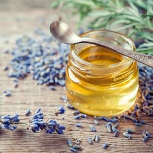 A small jar of lavender oil with tiny lavender flowers spread around the outside of a jar along with a few sprigs of fresh lavender.