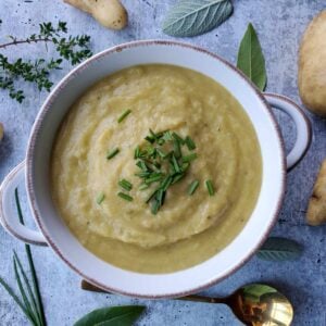 A white ceramic bowl with handles is full of creamy potato leak soup. It has been topped with fresh chopped chives and the bowl is surrounded by a few potatoes and fresh herbs.