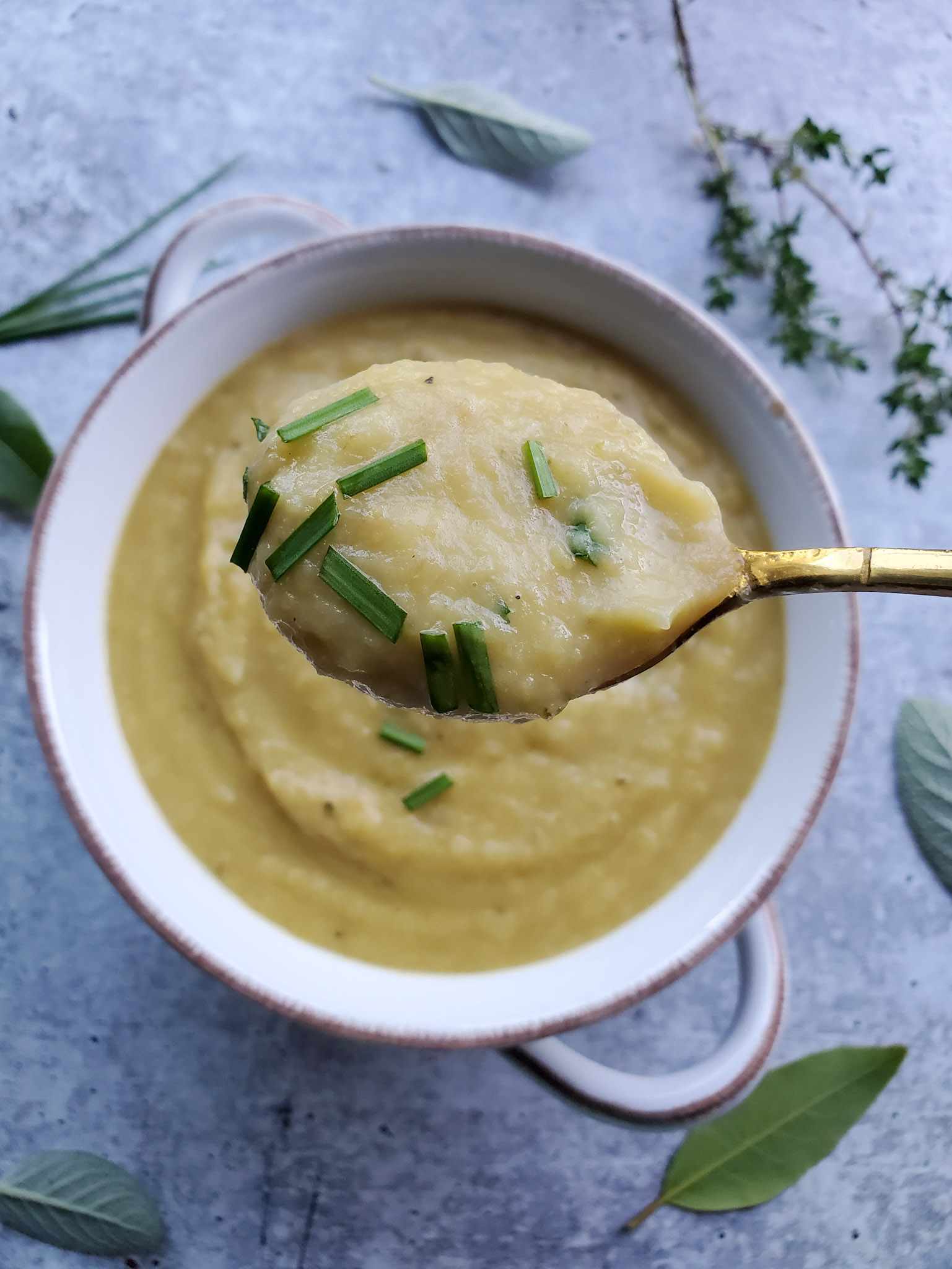 A close up image of a spoonful of potato leek soup that contains a couple pieces of fresh chives is shown. It is thick and golden beige in color. Below the spoonful lies a bowl of the soup with various fresh herbs arranged around the outside of the bowl. 