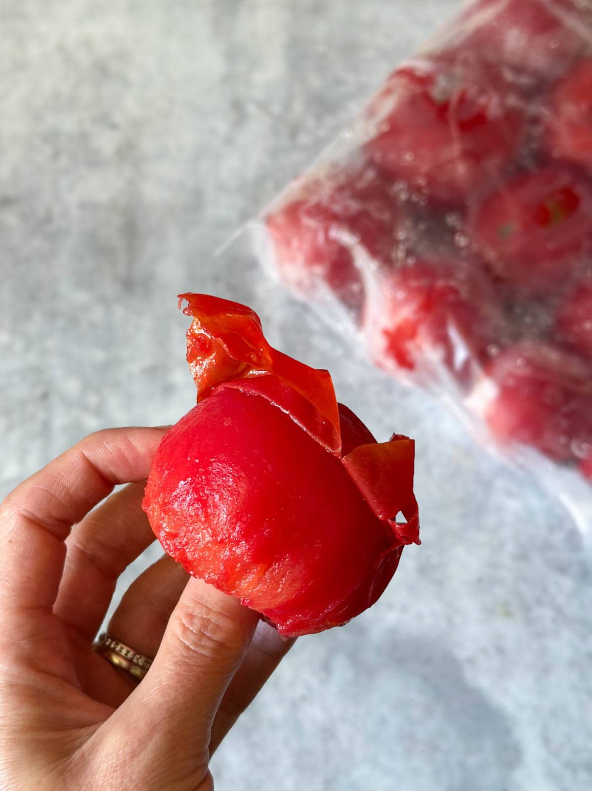 a hand holding a frozen, partially defrosted tomato with the skins hanging off of it, demonstrating how easily it peels