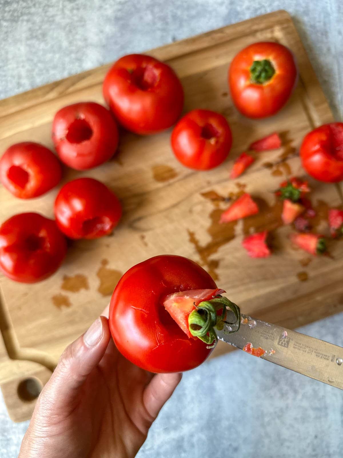 A hand holding a tomato, using a small paring knife to demonstrate cutting out the core and stem area. There is a cutting board and several whole cored tomatoes blurred in the background