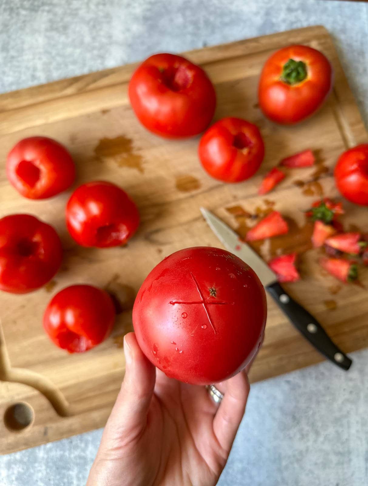 a hand holding a ripe tomato with a small X cut in the bottom of it, with a cutting board, knife, and cored whole tomatoes blurred in the background