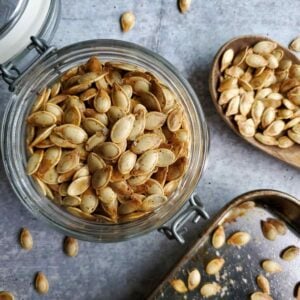 Roasted pumpkin seeds are inside a glass jar with a flip top lid. A wooden spoon and part of a baking sheet are nearby which roasted pumpkin seeds on them as well.